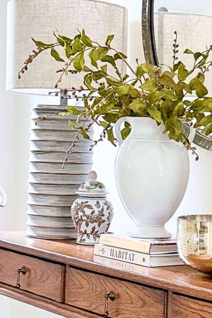 console table styled with tall textured lamp, white ceramic vase with greenery, stacked books, small patterned jar, and gold bowl for a collected look