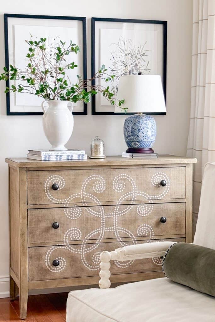 wood dresser styled with blue and white lamp, white vase with green branches, and framed botanical art in a calm, welcoming home