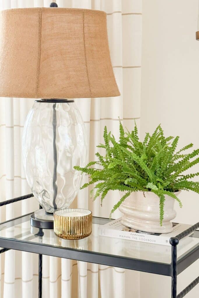 Winter vignette with a table lamp, potted fern, candle, and books styled on a glass side table for a fresh February home