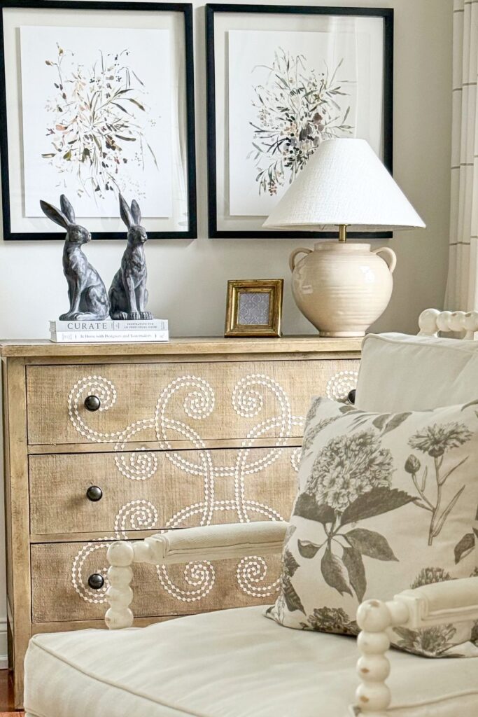 spring console table decorated with bunny figurines, stacked books, ceramic lamp, and botanical wall art