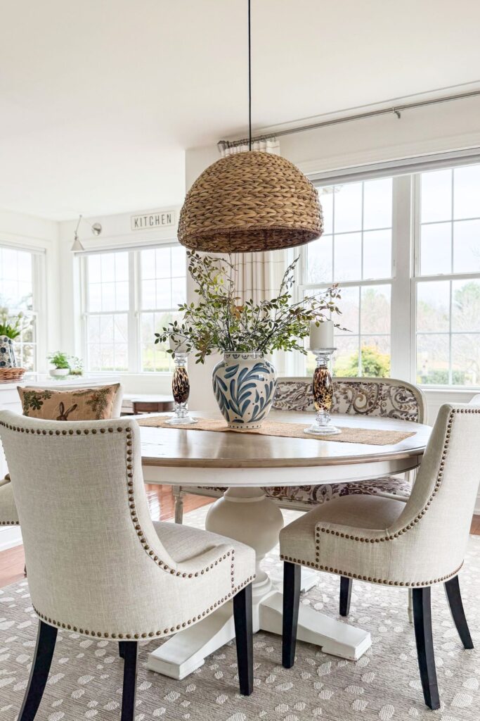 round dining table styled for spring with blue and white vase, greenery centerpiece, and woven pendant light above