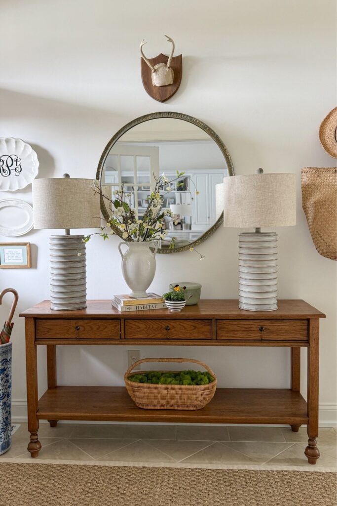 spring foyer console table styled with white vase of flowering branches, lamps, round mirror, and basket of moss