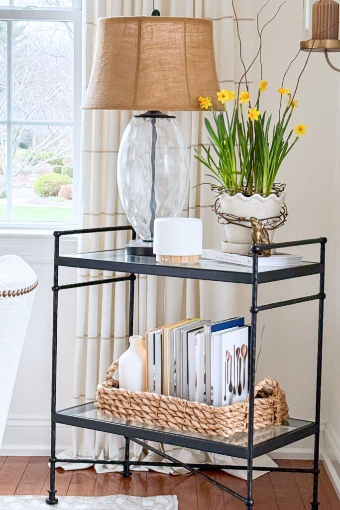 spring living room side table styled with a lamp, potted daffodils, books, and a basket in a bright neutral room