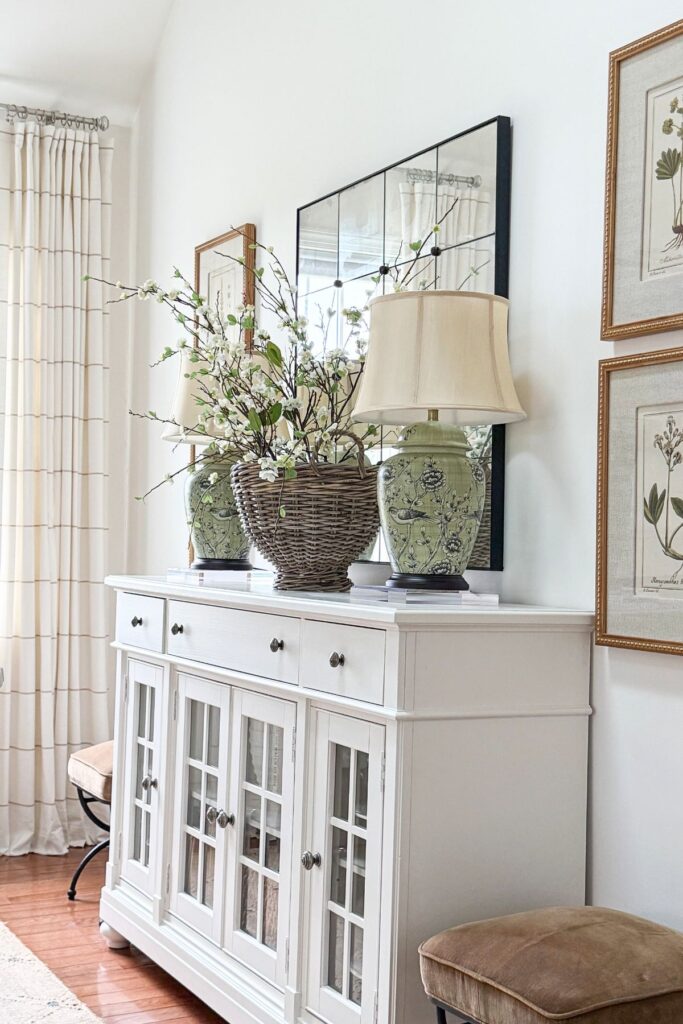 spring sideboard decorated with basket of flowering branches, green lamps, and botanical wall art