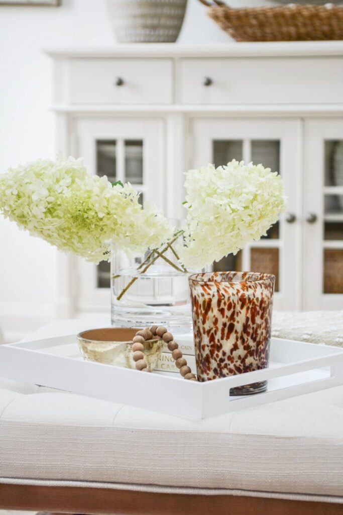 upholstered coffee table styled with white tray, hydrangeas, candle, and decorative accents in a neutral living room