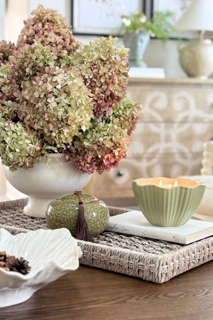 coffee table styled with hydrangeas in a white vase, woven tray, candle, and decorative accents in a neutral living room