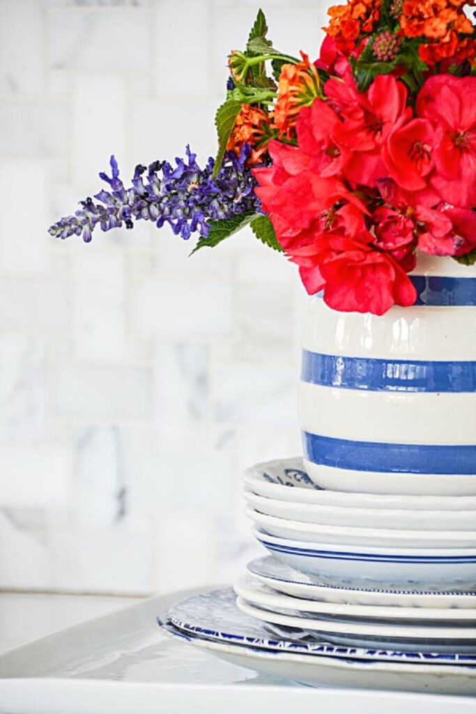 red and orange garden flowers with purple salvia in a blue and white striped crock on stacked blue and white plates for patriotic summer decorating