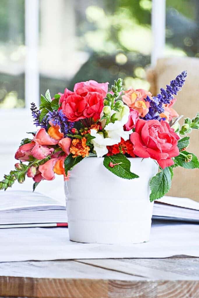 colorful summer flower arrangement with coral roses, lavender, and snapdragons in a white crock on a wooden table