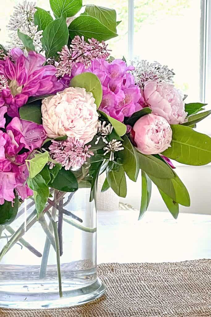 lush pink peony, azalea, and lilac flower arrangement in a clear glass vase on a jute rug near a bright window