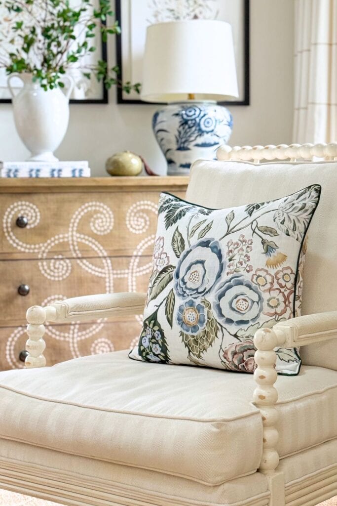 blue and pink floral throw pillow on a cream spindle chair with a blue and white ginger jar lamp and stenciled dresser in the background
