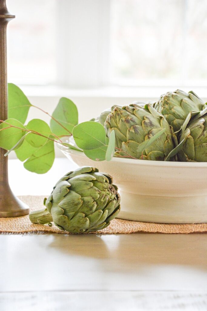 fresh artichokes in a white bowl with eucalyptus stems and a brass candlestick on a jute mat for a summer table centerpiece