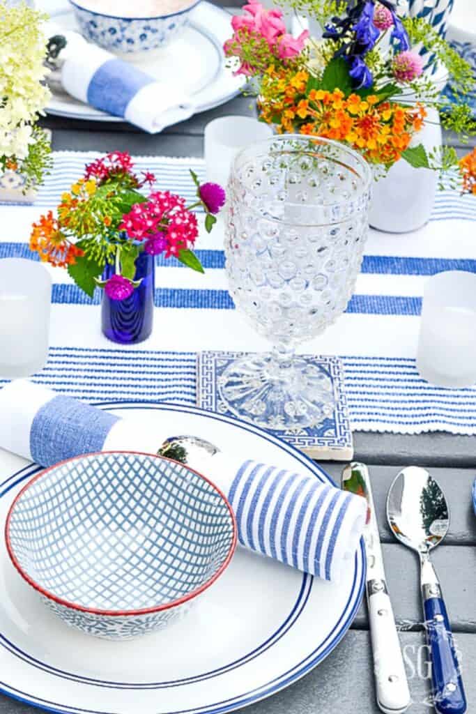 blue and white summer place setting with layered dishes, a hobnail glass, striped napkin, and small posies of colorful garden flowers in cobalt blue vases