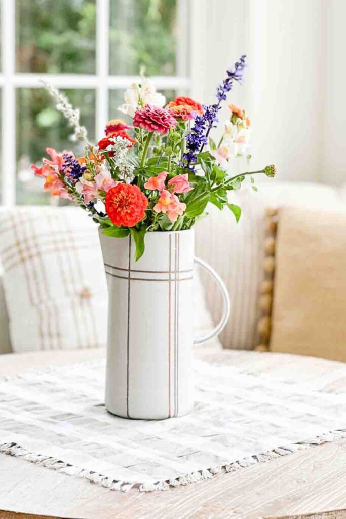 colorful summer garden flowers including zinnias, snapdragons, and salvia in a white grid pitcher on a fringed runner on a wood table