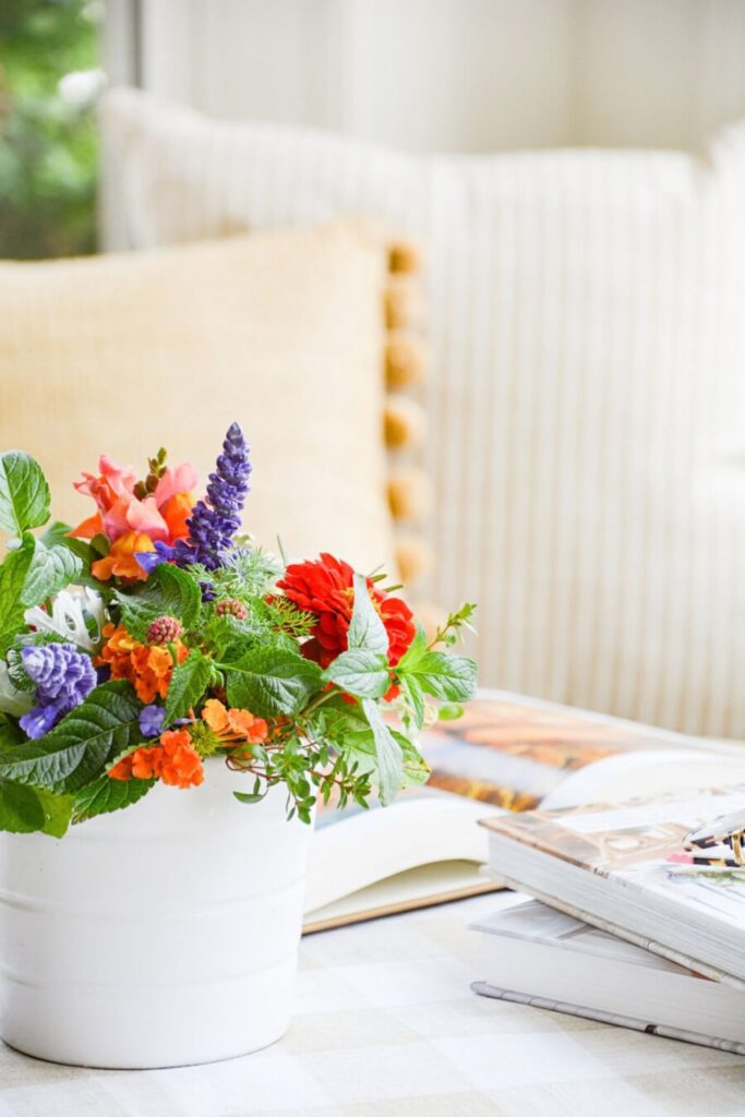 colorful summer garden flowers including zinnias, lupine, lantana, and snapdragons arranged in a white vase on a table with stacked books