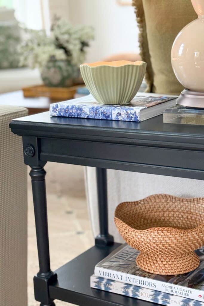 Small black end table styled with decorative bowls, stacked books, lamp, and shelf decor in a neutral living room