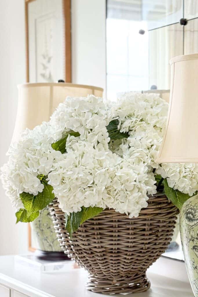 close up of faux white hydrangeas in a wicker basket next to a green chinoiserie bird lamp on a white sideboard with an antiqued mirror