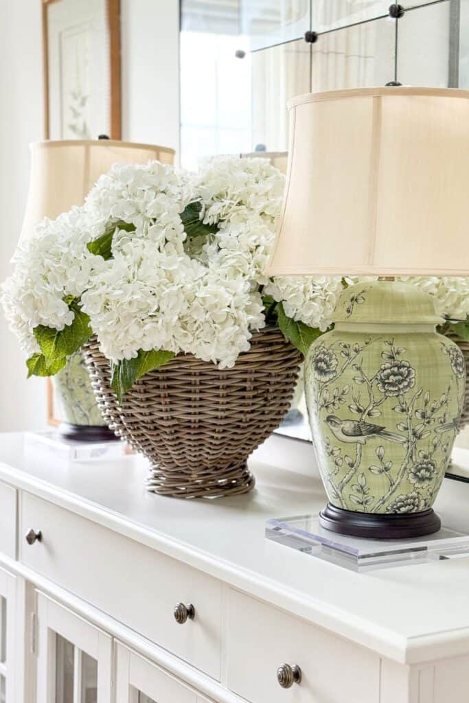 close up of faux white hydrangeas in a wicker basket next to a green chinoiserie bird lamp on a white sideboard with an antiqued mirror