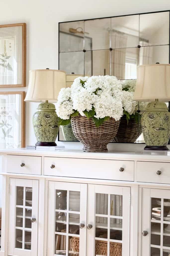 faux white hydrangeas in a wicker basket next to a green chinoiserie bird lamp on a white sideboard with an antiqued mirror