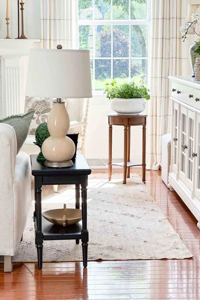 Small black end table in a neutral living room styled with a lamp, decorative accessory, and bowl on the lower shelf