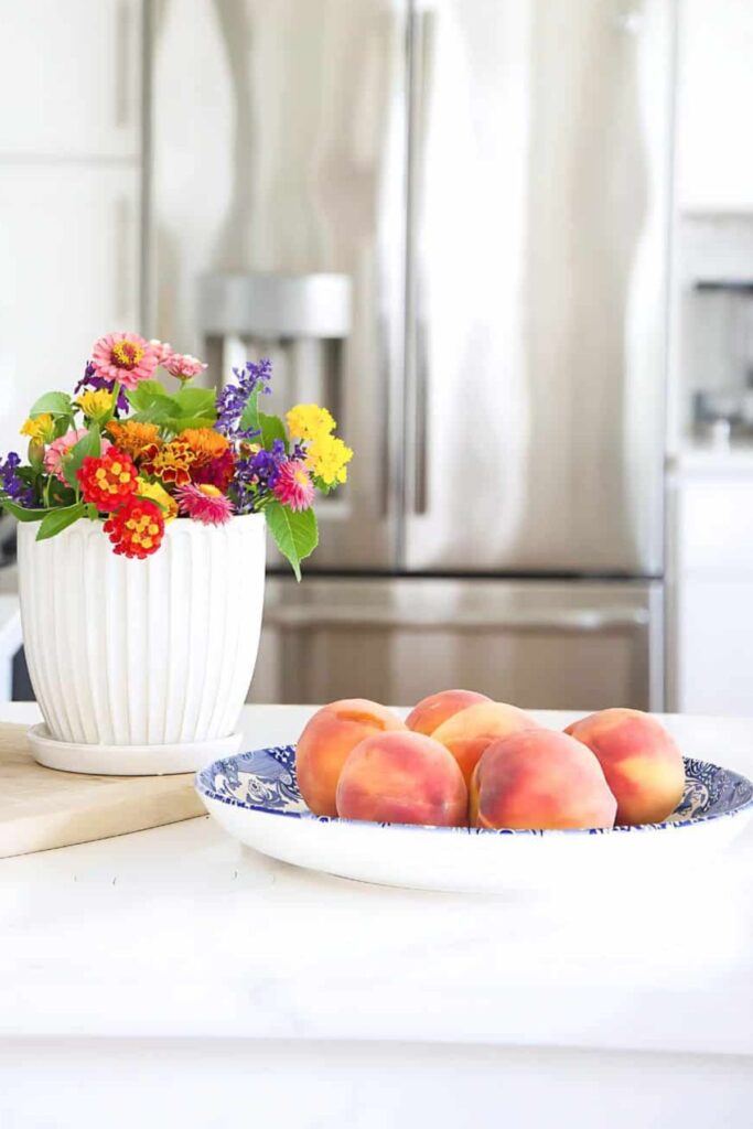 colorful garden flowers in a white ribbed pot and fresh peaches in a blue and white bowl on a white kitchen island