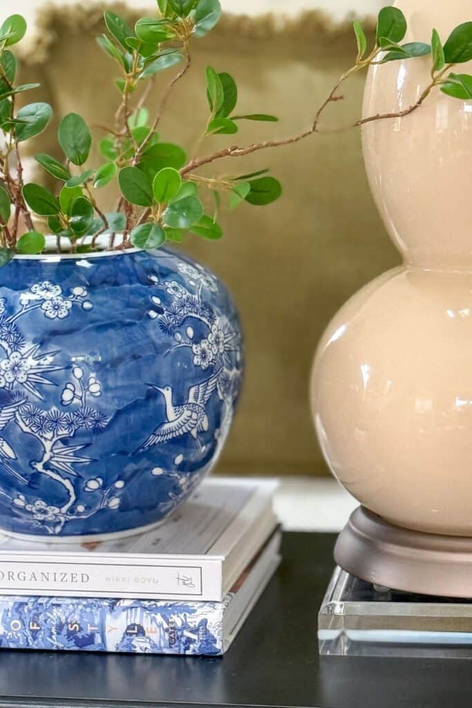 Layered decor on a small black end table with a blue and white vase, greenery, stacked books, lamp, and acrylic riser