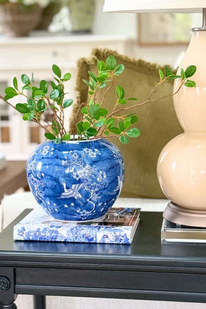 Small black end table decorated with a blue and white vase filled with greenery beside a lamp in a neutral living room