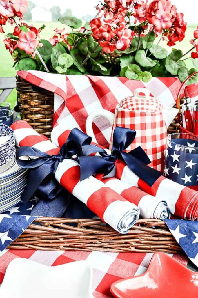 patriotic summer table setting with red white and blue napkins tied with navy ribbon, gingham ceramic jar, stars and stripes glassware, and red geraniums in a wicker basket