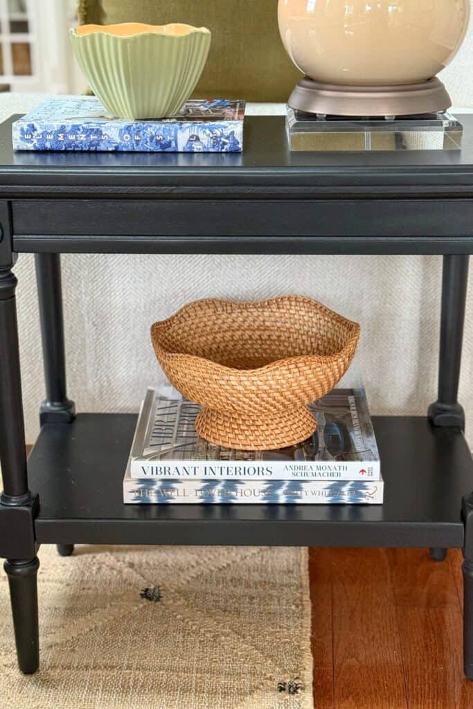 Small black end table with shelf styling using stacked books, woven bowl, lamp, and candle in a neutral living room