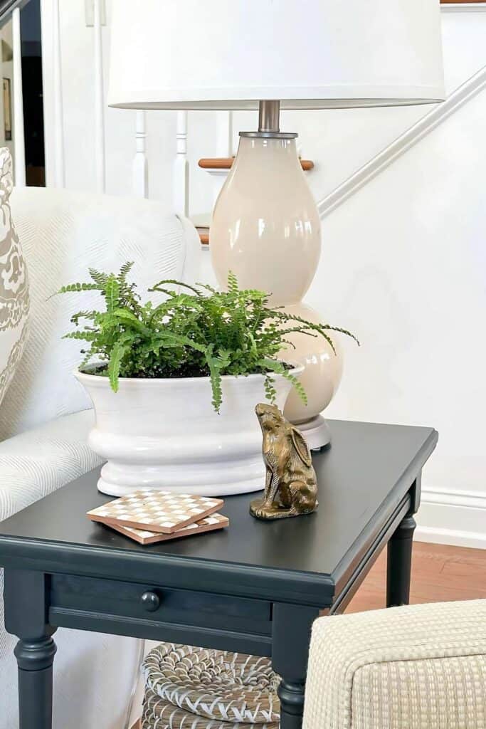 Small black end table decorated with a lamp, greenery, coasters, and basket shelf styling in a neutral living room