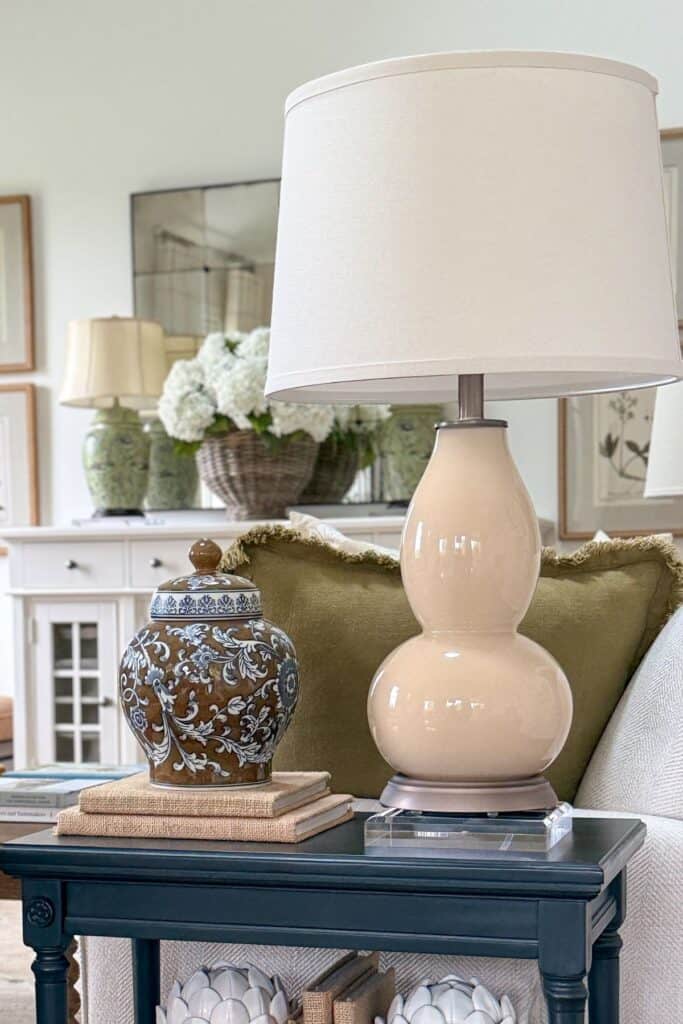 Small black end table styled with stacked neutral books, decorative ginger jar, lamp, and acrylic riser in a cozy living room