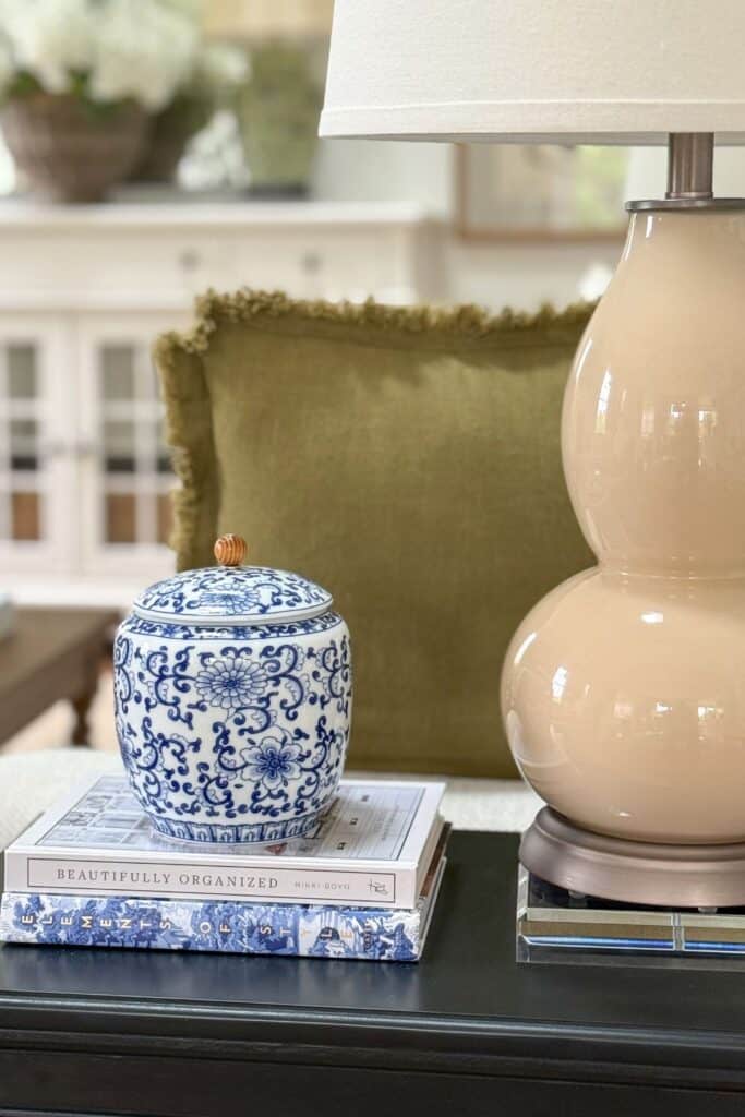 Small black end table styled with stacked books, blue and white ginger jar, lamp, and acrylic riser in a neutral living room