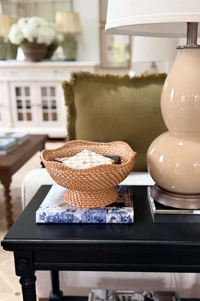 Small black end table styled with a woven bowl, stacked books, lamp, and acrylic riser in a neutral living room