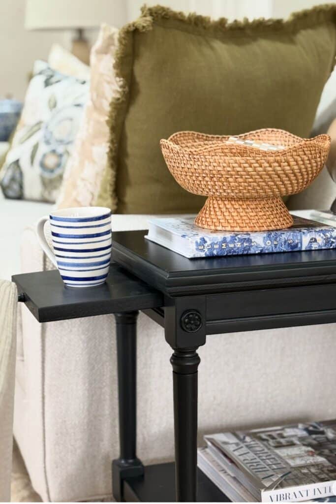 Small black end table with a pull out tray, woven bowl, books, and striped mug beside a neutral chair