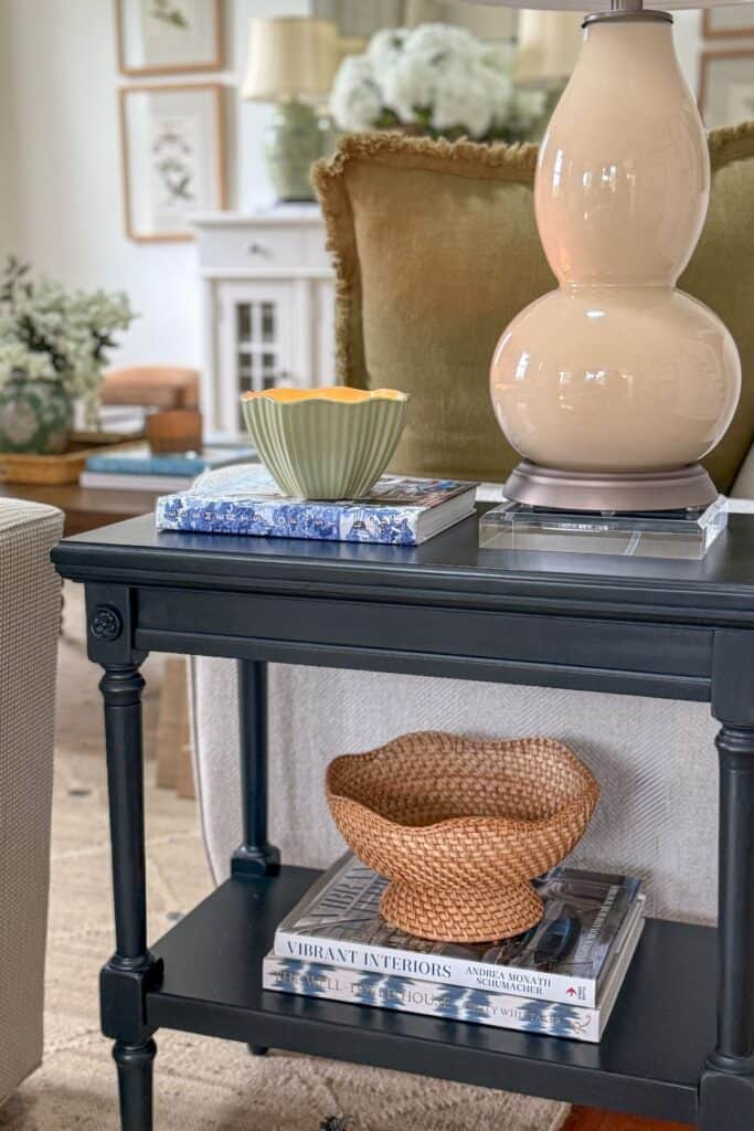 Small black end table styled with a lamp, candle, books, and woven bowl on the lower shelf in a neutral living room