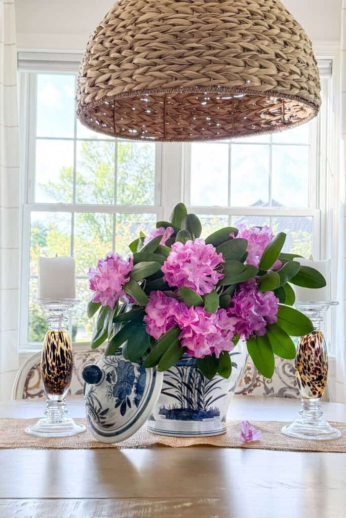spring dining table centerpiece with pink flowers in blue and white pitcher, woven pendant light, and glass candlesticks