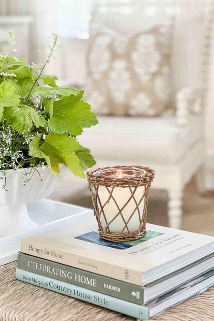 wicker wrapped candle on a stack of home decor books next to a white vase of fresh green leaves and wildflowers on a woven coffee table
