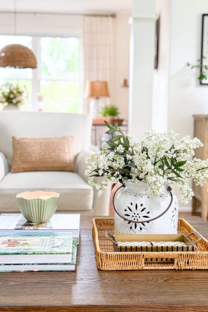 summer coffee table vignette with white flowers and eucalyptus in a white pierced ceramic pot on a wicker tray with stacked books and a green candle bowl