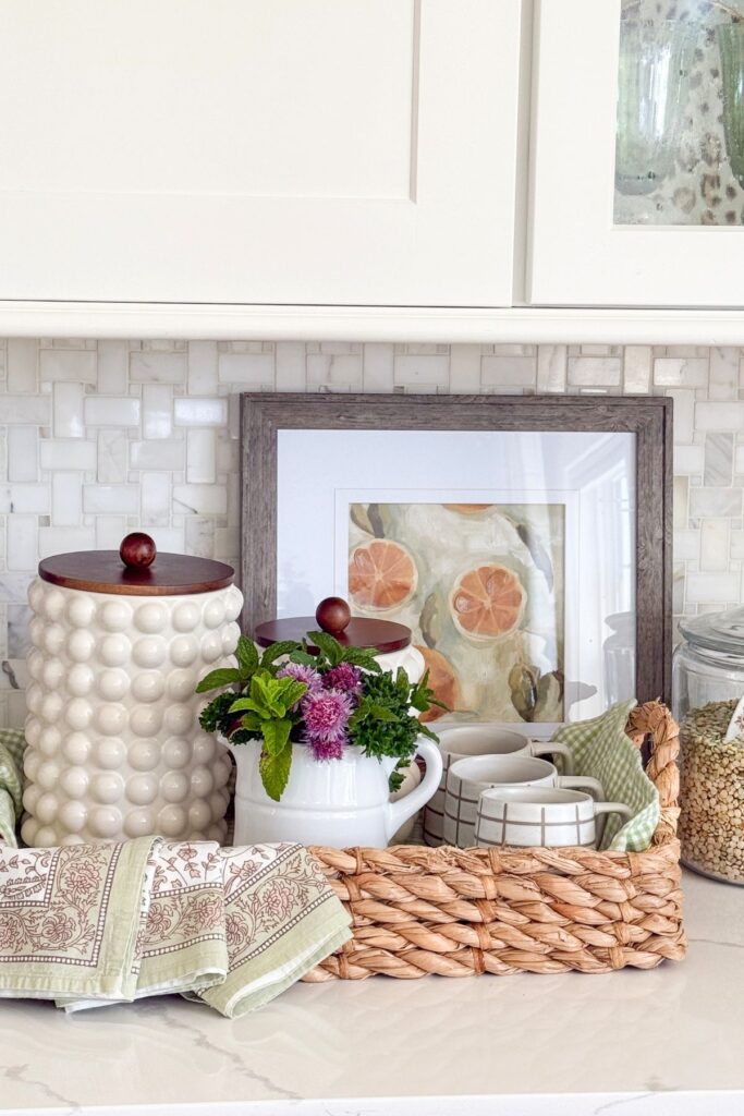 summer kitchen counter coffee bar vignette with a white bobble canister, fresh herbs and chive blossoms in a white pitcher, grid mugs in a woven basket tray, floral linen napkins, and a framed citrus print on a marble backsplash