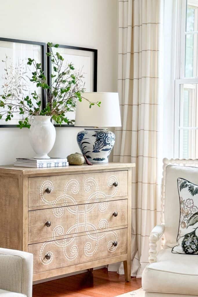 summer room vignette with a floral throw pillow on a cream spindle chair, white urn with green branches, blue and white ginger jar lamp, and botanical prints on a stenciled dresser
