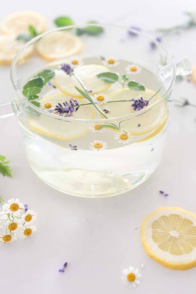 angled view of a glass simmer pot filled with lemon slices, lavender sprigs, fresh mint, and chamomile flowers on a white surface