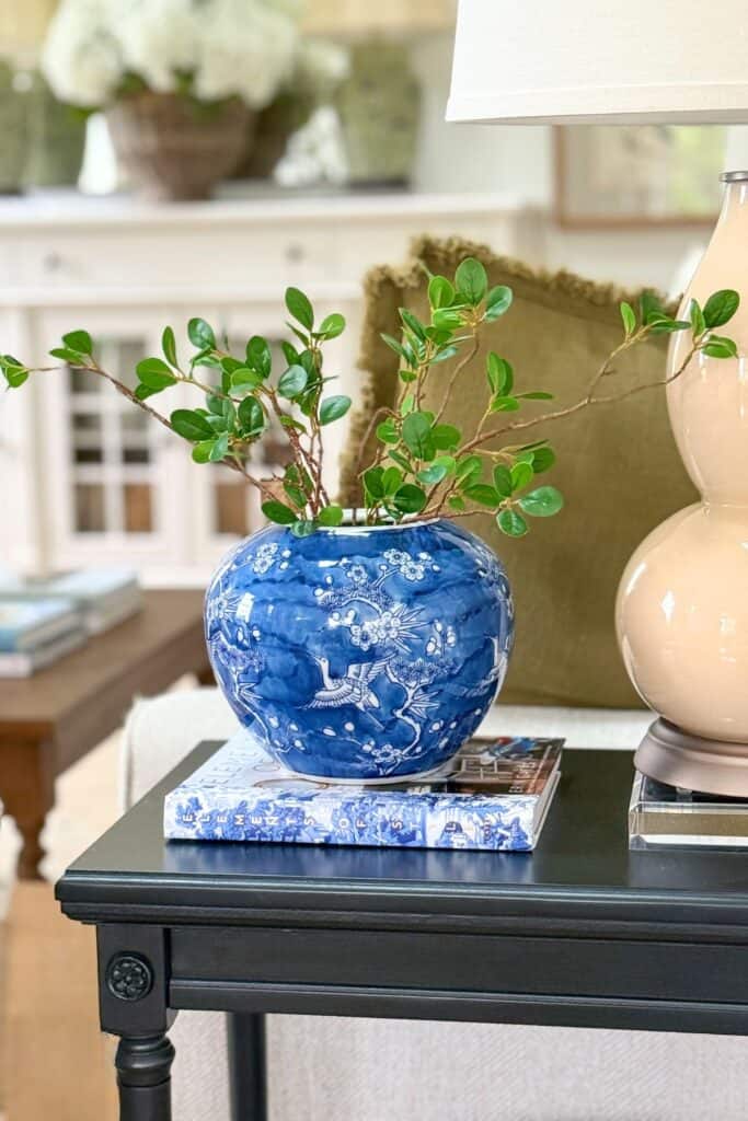 Small black end table decorated with a blue and white vase, greenery, books, and lamp in a neutral living room