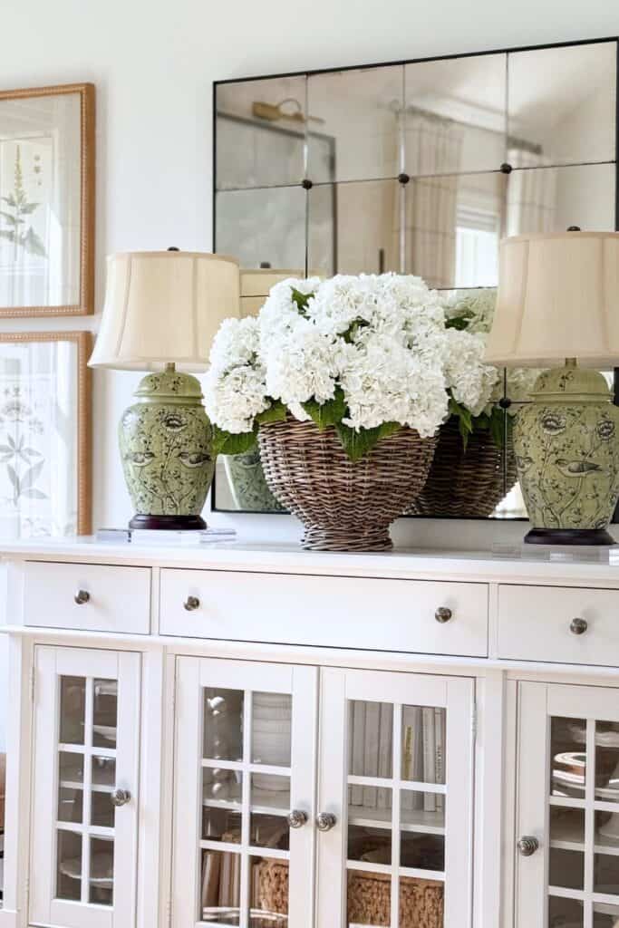 white hydrangeas in a wicker basket on a white buffet flanked by green ginger jar lamps in front of a grid mirror for summer decorating