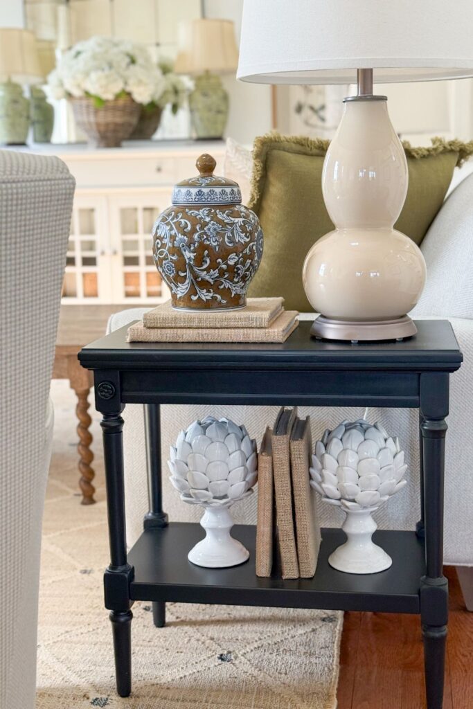 A neutral cream double gourd ceramic table lamp on a black end table styled with burlap wrapped books, a brown and blue ginger jar, and white artichoke finials
