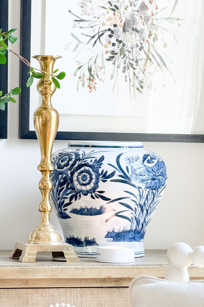 A tall brass candlestick standing beside a large blue and white chinoiserie ginger jar with a small puck light on a wood console table, showing the materials for a DIY lamp.