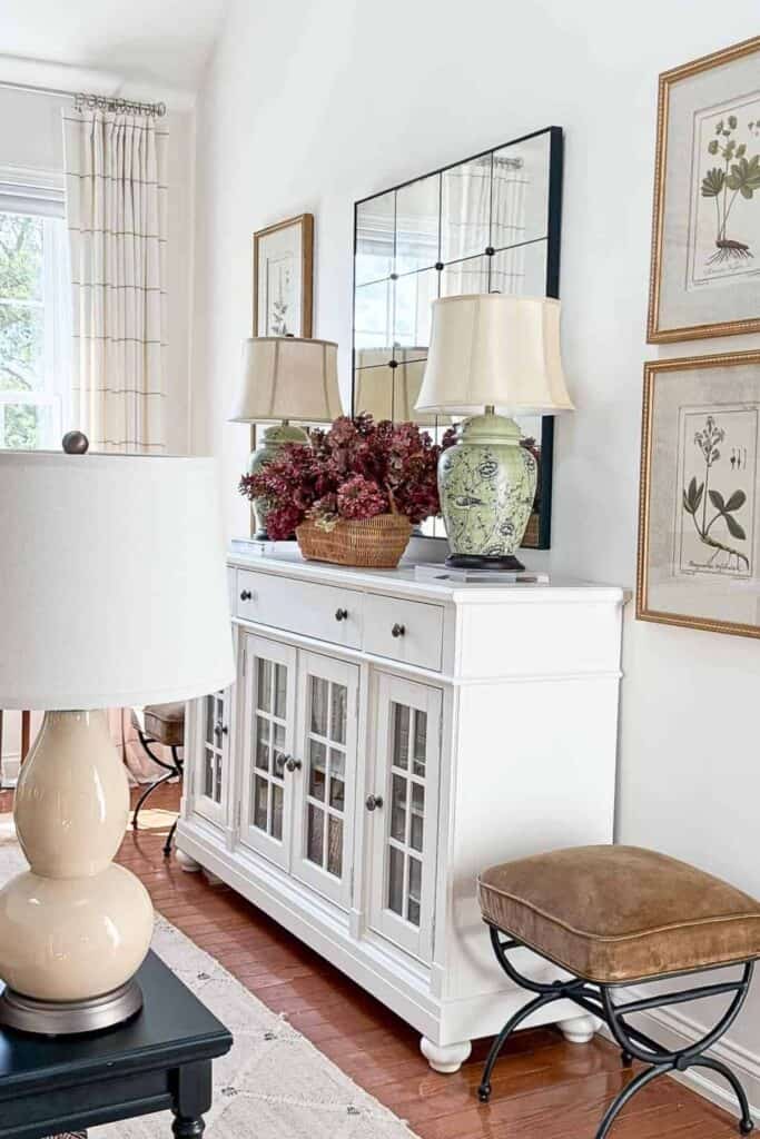 A pair of green ceramic ginger jar table lamps on a white buffet styled with a wicker basket of burgundy florals, a large mirror, and botanical art in a living room