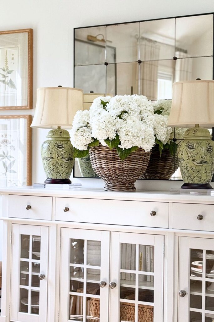 A pair of green ceramic ginger jar table lamps on a white buffet flanking a large wicker basket of white hydrangeas with a large mirror behind
