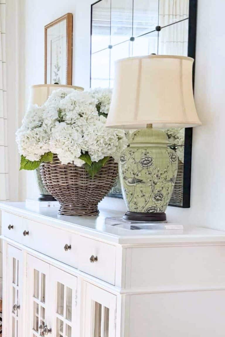 A green ceramic ginger jar table lamp on a clear acrylic riser on a white buffet next to a large wicker basket of white hydrangeas with a large mirror behind