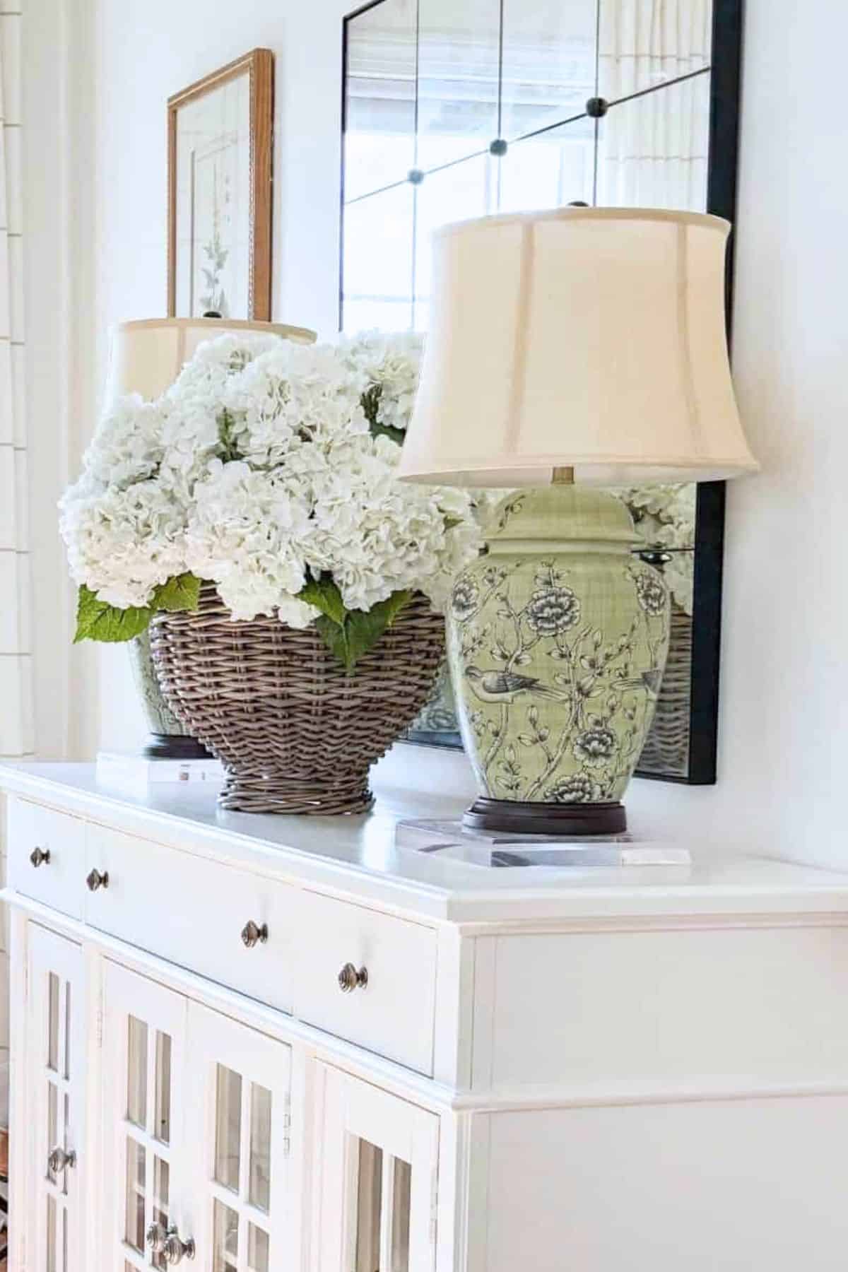 A green ceramic ginger jar table lamp on a clear acrylic riser on a white buffet next to a large wicker basket of white hydrangeas with a large mirror behind