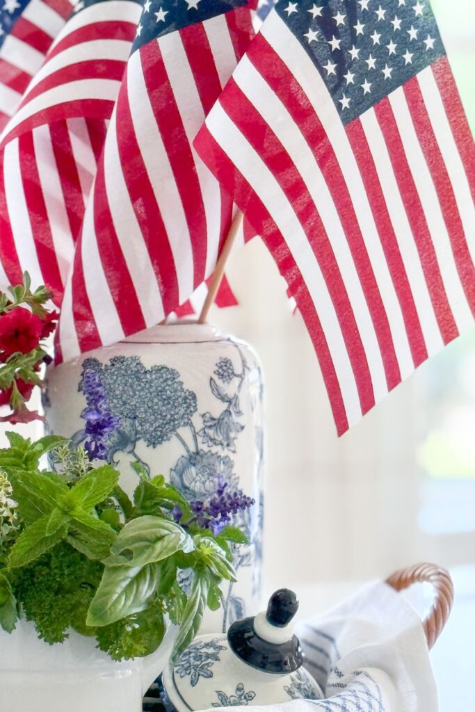 American flags standing in a tall blue and white chinoiserie ginger jar beside fresh garden herbs in a white pitcher and a small blue and white teapot on a wicker tray in a patriotic kitchen island vignette.