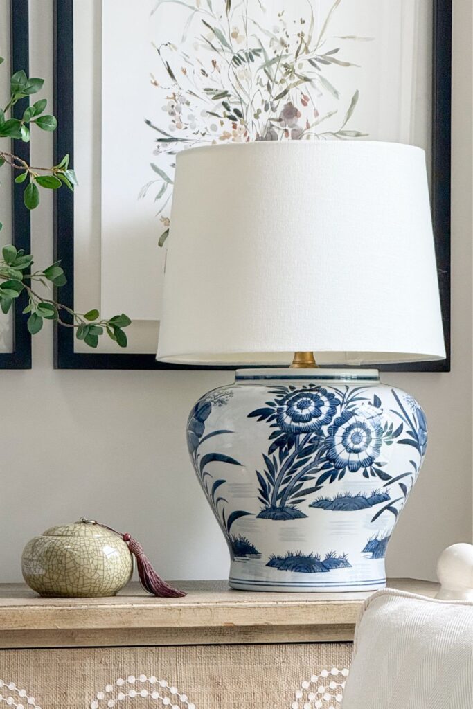 A large blue and white ceramic ginger jar table lamp on a natural wood console with framed watercolor botanical art and a small green ceramic decorative box